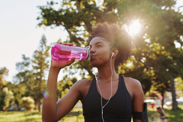 Fitness athlete young african american woman listening to music on earphones drinking water in a reusable water bottle after working out