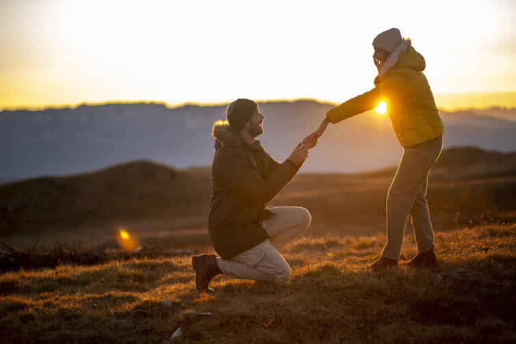 Silhouettes of a man making a marriage proposal to his girlfriend on the mountain peak at sunset.