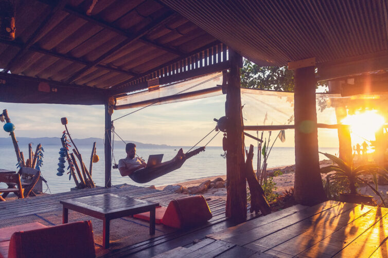 Man laying in hammock with a laptop by the beach at sunset 