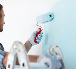 Man standing on a ladder painting a wall blue with a paint roller.