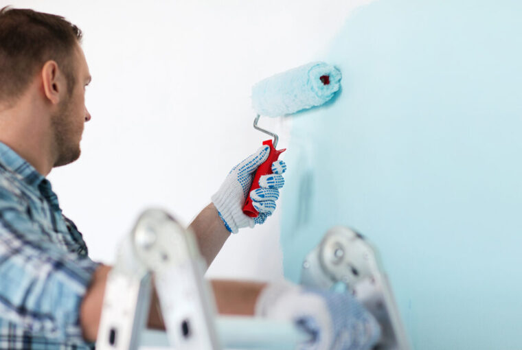 Man standing on a ladder painting a wall blue with a paint roller.