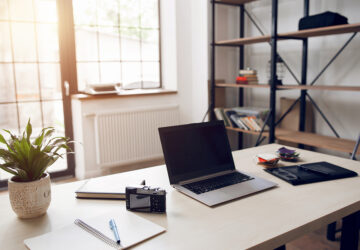 Home office with large windows, desk, shelving and laptop.