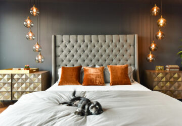 Bedroom with dark painted walls, cosy bed with small black and white dog sleeping. Stylish glass hanging pendent lights.