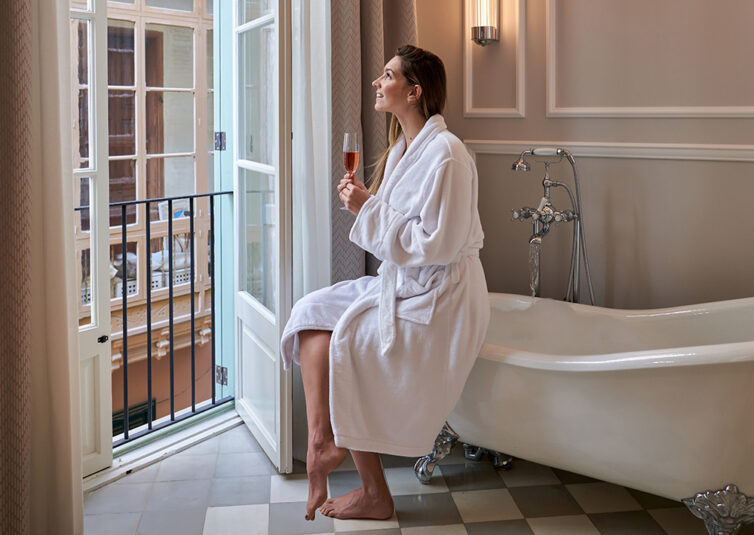 Women in white dressing gown sat on freestanding bath, holding glass of champagne