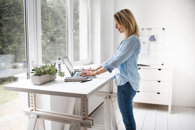 Women with blond hair wearing jeans and blue blouse standing by  large window and white egonomic standing desk