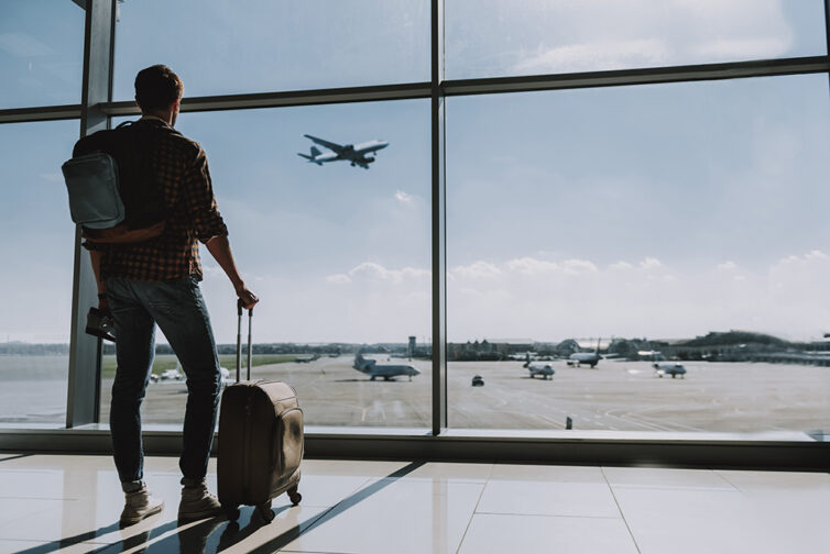 Man with suitcase on wheels standing at large window in airport looking at airplans