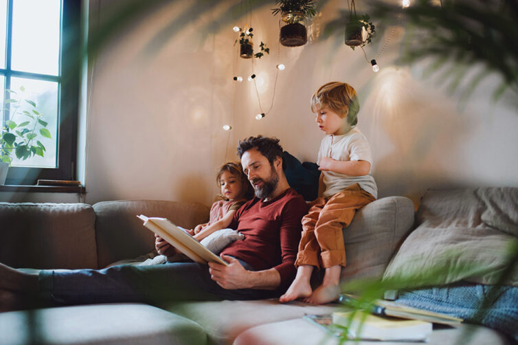 Dad, daughter and son sat on sofa. Fairy lights on the wall.