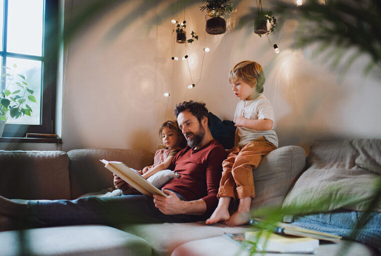 Dad, daughter and son sat on sofa. Fairy lights on the wall.