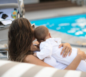 Women holding a baby in a white romper suit whilst on holiday next to a swimming pool.