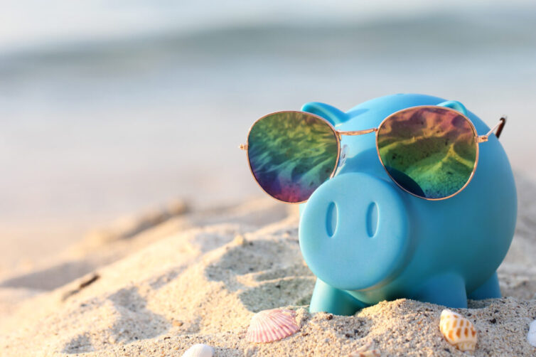 Blue Piggy Bank placed on sand with sunglasses on. Saving for a holiday