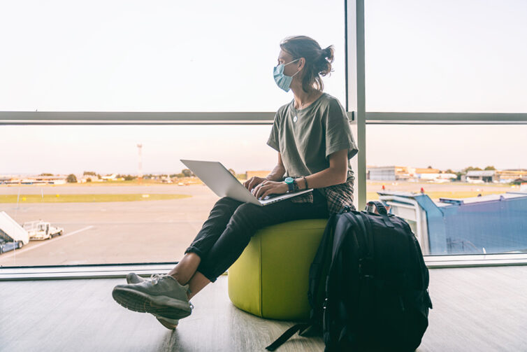 Women sate on green seat at airport with bakpack next to her. Women wearing facemask, whilst using laptop.